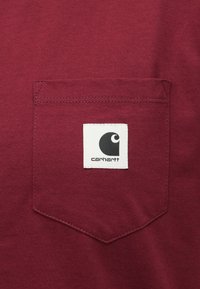 T-shirt in cotone color bordeaux con taschino decorato da cuciture bianche e un'etichetta quadrata in bianco e nero sulla tasca.