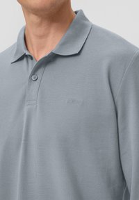 Mann trägt ein hellblaues kurzärmliges Poloshirt mit Kragen und zwei Knöpfen, das ein dezentes besticktes Markenlogo auf der Brust aufweist.