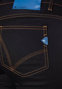 Czarne jeansy z denimu z kontrastującym pomarańczowym przeszyciem i niebieską skórzaną metką z logo. Posiadają małą kieszonkę o charakterystycznym kształcie.