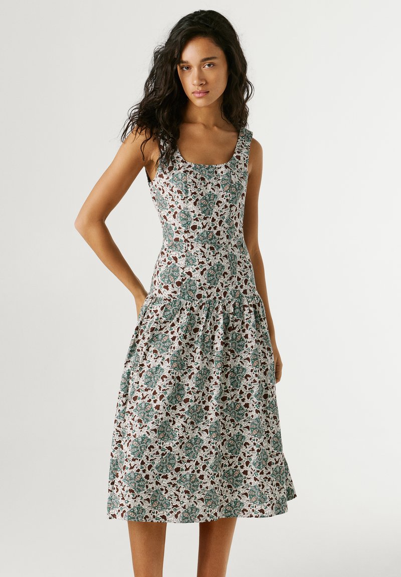 Mujer con vestido sin mangas de longitud media con estampado floral verde y marrón, de pie con una mano en la cadera contra un fondo liso.