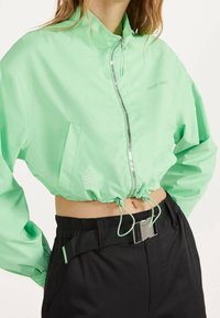 Mintgrön kort bomberjacka med silverfärgad dragkedja, elastisk nederkant och logotypapplikation. Matchad med svarta byxor som har ett bälte.