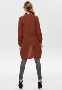 Cárdigan marrón de punto con textura de canalé, mangas largas y espalda abierta. Combinado con jeans ajustados grises y zapatos de tacón negro.