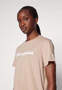 Beige īsām piedurknēm T-kreklam ar balto un rozā "Champion" logotipu. Izgatavots no mīkstas auduma ar brīvu piegulšanu; logotips uz krūšu partijas un piedurknes.
