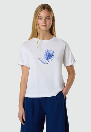 Junge Frau mit langen blonden Haaren trägt ein weißes T-Shirt mit blauem Korallen- und Muschel-Print, kombiniert mit dunkelblauen Shorts, stehend vor einem schlichten Hintergrund.