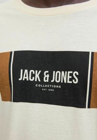 Kreemist T-särgist, millel on pruun ja must ristkülikukujuline graafika, mis sisaldab "JACK & JONES" teksti ja "COLLECTIONS EST. 1990" julge valge fondiga.
