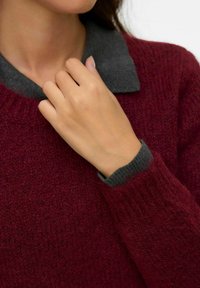 Burgunderfarbener Pullover mit strukturiertem Finish, ausgestattet mit einem grauen Kragen und gerippten Bündchen, teilweise gezeigt mit einer Hand, die den Kragen berührt.