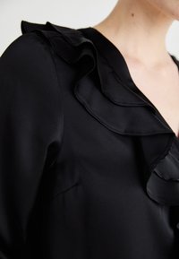 Blusa nera con scollatura e maniche a volant, realizzata in tessuto liscio, con un design aderente e una leggera brillantezza.
