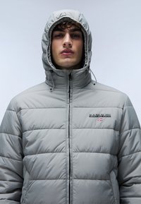 Lys grå pufferjakke med hætte, med et quiltet design, frontlynlås og et logo på brystet. Glat, skinnende materiale.