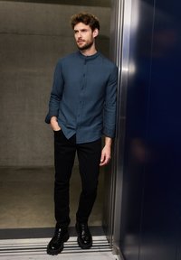 Koszula z długim rękawem, zapinana na guziki, w ciemnym odcieniu teal, noszona z dopasowanymi czarnymi spodniami i czarnymi butami na sznurówki, oparta o windę.