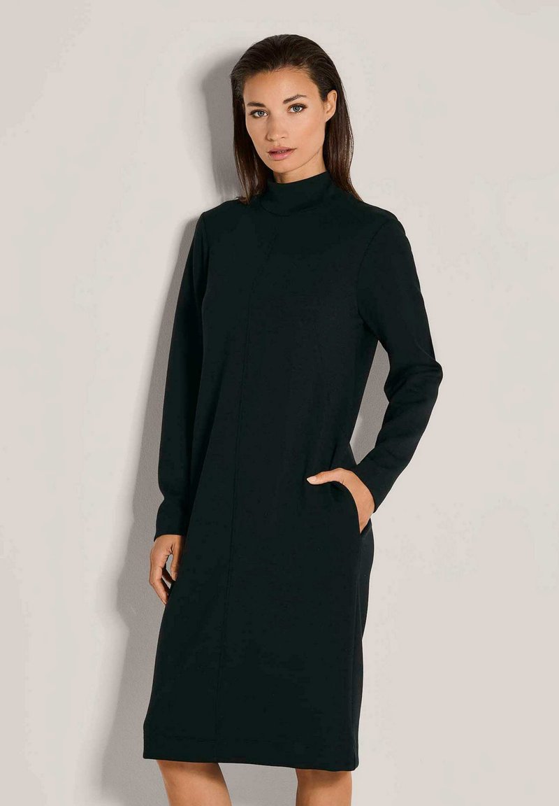 Frau steht in einem langärmeligen schwarzen Midikleid mit hohem Kragen und hat die rechte Hand in der Seitentasche.