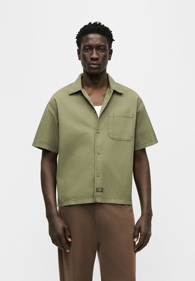 Hombre de pie, vistiendo una camisa de manga corta color verde oliva abotonada sobre una camiseta blanca y pantalones marrones, con un fondo liso.