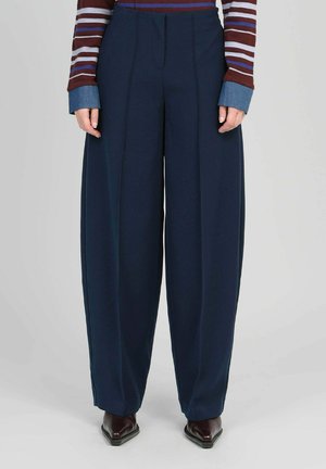 Personne portant un pantalon large bleu marine, un haut à manches longues bordeaux rayé avec des poignets bleus, et des chaussures sombres à bout pointu, debout devant un fond uni.
