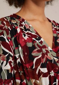 Stoff mit Blumenmuster, der rote, grüne und weiße Blumen auf schwarzem Hintergrund zeigt. V-Ausschnitt-Design mit Raffungen am Halsauschnitt.