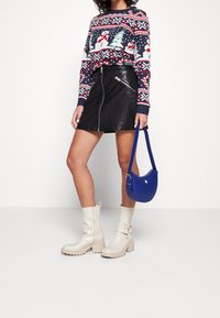 Femme portant un pull festif motif bonhomme de neige, jupe en cuir noire, bottes blanches, tenant un sac à bandoulière bleu, debout devant un fond blanc.