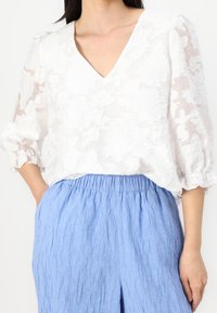 Vit blus med V-ringning, texturerat blommönster och puffärmar, kombinerad med ljusblå texturerade byxor.