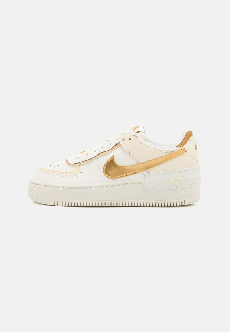 Baskets blanches basses avec le swoosh Nike doré, semelle blanche et zone perforée au niveau des orteils, présentées de profil sur fond uni.