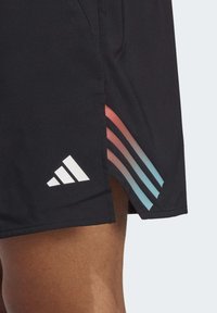 Bermudas deportivas negras con un logo de Adidas en blanco, diseño de tres rayas en degradado en azul y rosa, con aberturas laterales para mayor movilidad.
