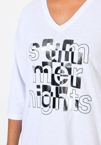 Witte V-hals T-shirt met een zilverkleurige metallic grafische print die "zomernachten" in variërende lettertypen en maten weergeeft. 3/4-lange mouwen.