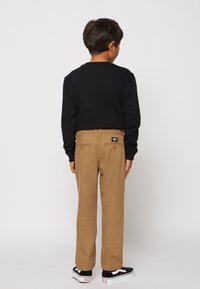 Zwarte langemouwen shirt, beige broek met achterzakken en zwart-witte sneakers. De broek heeft een label op de taille en een rechte pasvorm.