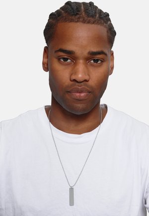 Collier en argent avec un pendentif en forme de tag rectangulaire sur une fine chaîne, porté par une personne en t-shirt blanc uni avec des cheveux tressés.