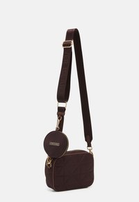 Hnědá prošívaná crossbody taška se zlatými doplňky a připojenou kulatou mincovkou s logem. Nastavitelný tkaninový popruh pro pohodlí.