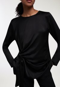 Top lungo manica in satin nero con dettaglio di nodo intrecciato in vita e scollatura rotonda, caratterizzato da un tessuto liscio e fluido con una consistenza morbida.