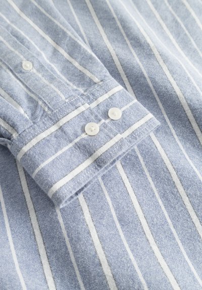 Chemise rayée bleu clair avec des rayures blanches verticales, deux boutons blancs sur le poignet et une finition en tissu doux et texturé.