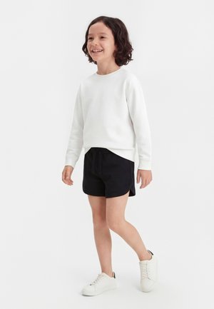 Enfant aux cheveux bouclés souriant, marchant, portant un haut blanc à manches longues, un short noir et des baskets blanches sur fond uni.