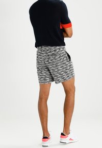 Homme debout les bras croisés, portant une chemise à manches courtes noire et rouge, un short grise texturé et des baskets blanches avec des accents roses.