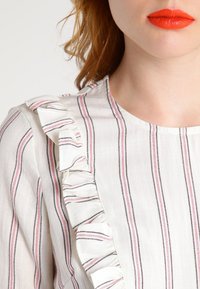 Camicia in tessuto bianco con righe verticali rosse e nere, caratterizzata da un dettaglio arricciato sulle spalle. Texture liscia, scollatura rotonda.