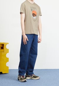 Beige t-shirt med NASA-grafik, blå denimjeans og flerfarvede sneakers. Placeret ved siden af en gul opbevaringsenhed på en blå gulv.