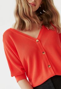Czerwony dziergany kardigan z dekoltem w serek, krótkimi rękawami i trzema drewnianymi guzikami z przodu; lekka, fakturowana tkanina.