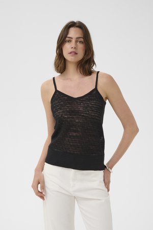 Žena s hnedými vlasmi po plecia, oblečená v čiernom textúrovanom tielku bez rukávov a bielych nohaviciach, stojaca pred jednoduchým pozadím.