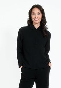 Juoda zip-up palaidinė su apykakle, ilgomis rankovėmis ir lygiu paviršiumi. Suderinta su juodomis kelnėmis, sukuriant vientisą, minimalistinį įvaizdį.