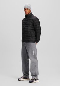 Zwarte pufferjack met horizontale stiksels, hoge kraag, twee met rits afgesloten zijvakjes, gecombineerd met grijze jeans en multicolor sneakers.