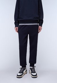 Person trägt schwarze Jogginghose, schwarzen Hoodie über einem grauen Shirt und weiße und schwarze Turnschuhe und steht vor einem hellgrauen Hintergrund.