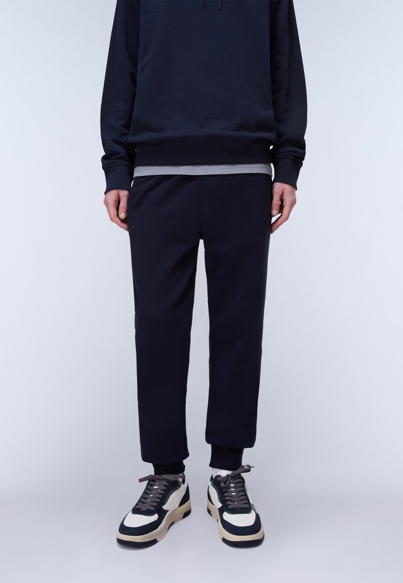 Person trägt schwarze Jogginghose, schwarzen Hoodie über einem grauen Shirt und weiße und schwarze Turnschuhe und steht vor einem hellgrauen Hintergrund.