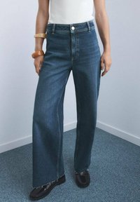 Wijdelopende denimjeans in een donkere wasbeurt met een ruwe zoom, met vijf zakken en een knoopsluiting, gecombineerd met zwarte schoenen en een witte top.