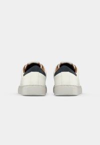 Paire de baskets blanches basses avec languettes de talon noires et semelles en caoutchouc gris clair, vues de dos sur un fond uni.