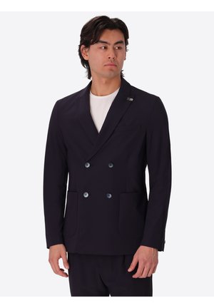 Uomo con blazer doppiopetto nero sopra una camicia bianca, in piedi e che guarda di lato su uno sfondo bianco uniforme.