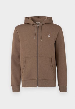 Smeđa zip-up dukserica od mekog materijala s prednjim džepom, podesivim vezicama i malim belim logotipom na grudima.