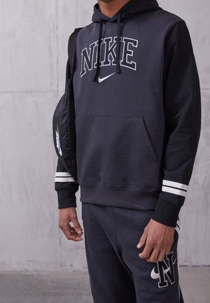 Person trägt schwarzen Nike-Hoodie und passende Jogginghose mit weißem Logo, trägt eine schwarze Nike-Umhängetasche vor grauem Wandhintergrund.