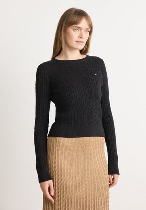 Svart strikket genser med kabelmønster, rund hals og ribbestrikkede mansjetter, parret med et strukturert beige skjørt. Enkel logoaccent på genseren.