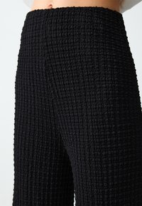 Des pantalons larges noirs texturés présentent une surface froncée et une taille haute élastique. Le matériau semble doux et respirant.