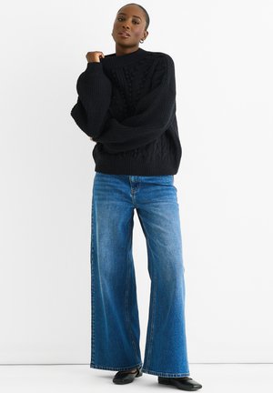 Black cable-knit sweater with high neckline, oversized sleeves. Paired with wide-leg blue jeans and black shoes. Solid colors, textured patterns.