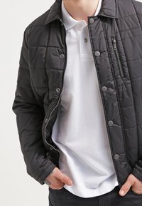 Man i en svart vadderad jacka över en vit polo skjorta med händerna i fickorna på svarta byxor, som visar en liten handtatuering.