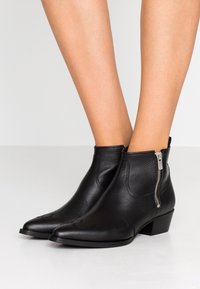 Botas de cuero negras hasta el tobillo con puntera afilada, costuras decorativas y cremallera lateral. Tacón bajo y textura suave.