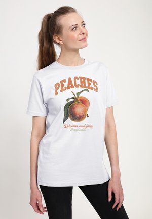 Bílé bavlněné tričko s grafikou broskve a nápisem "PEACHES" a "Delicious and juicy" v barevném tisku. Modelka má na sobě černé džíny.