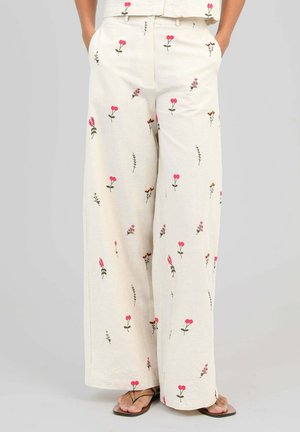 Pantalon crème à jambes larges avec de petites impressions florales roses et marron dispersées, porté avec un haut assorti et des sandales.