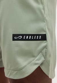 Shorts vert clair avec une étiquette noire affichant le mot « ENDLESS » en blanc, portés par une personne à la peau foncée visible.
